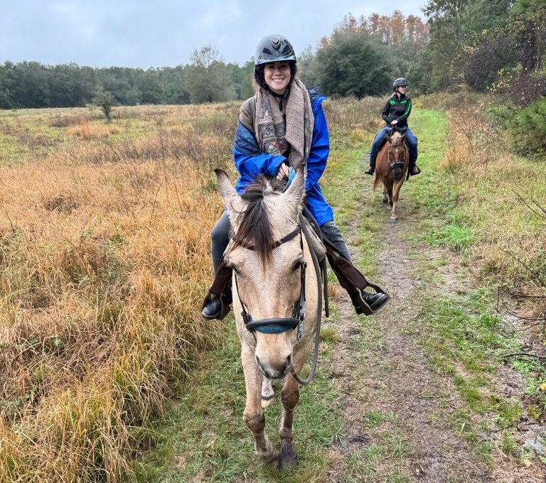 Two people horseback riding on a grassy trail through a field, one in front, both wearing helmets.