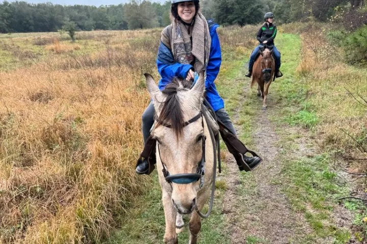Two people horseback riding on a grassy trail through a field, one in front, both wearing helmets.