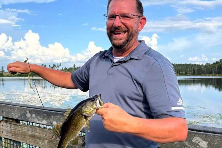 Man holding a fish on a dock by a lake with a cloudy sky.
